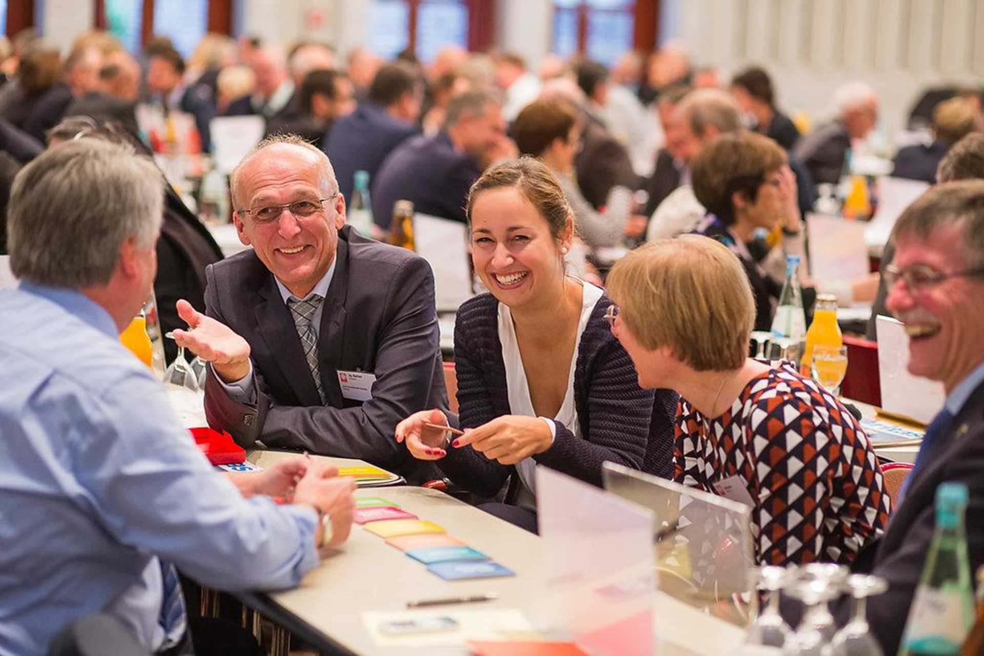 Gruppengespräch bei einer Konferenz mit mehreren Personen, die lächeln und interagieren. Auf dem Tisch liegen bunte Karten und Getränke.